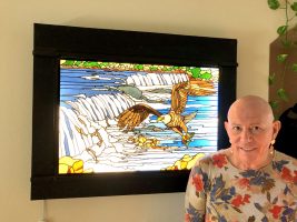 Lee Richards with “The Eagle at the Falls,” one of his stained-glass works mounted on a light box. (© FlaglerLive)