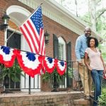 A couple poses in front of a brick home decorated with the American flag and bunting. Homeownership, often considered key to the American Dream, is difficult to achieve for many families due to rising housing costs. Kingfisher Productions via Getty Images