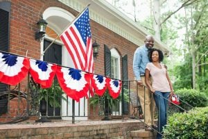 A couple poses in front of a brick home decorated with the American flag and bunting. Homeownership, often considered key to the American Dream, is difficult to achieve for many families due to rising housing costs. Kingfisher Productions via Getty Images