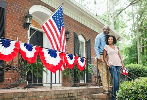 A couple poses in front of a brick home decorated with the American flag and bunting. Homeownership, often considered key to the American Dream, is difficult to achieve for many families due to rising housing costs. Kingfisher Productions via Getty Images