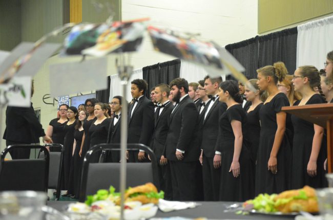 Amy Fulmer's Formality Singers opened the symposium with a performance. At every table, a parasol-like arrangement of pictures illustrated every school's flagship program. (© FlaglerLive)