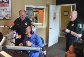 County Commissioner Dave Sullivan being arrested this morning near the end of Free For All Friday as it aired on WNZF, with Sheriff Rick Staly serving the warrant and reading it on the air, Chief Mark Strobridge preparing to handcuff the commissioner, and Relay For Life's Judy Mazzella in the lower right corner. Click on the image for larger view. (© FlaglerLive), 