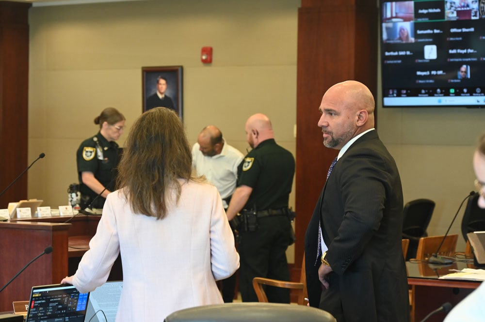 Defense attorney Aaron Delgado, right, speaks with Assistant State Attorney Melissa Clark as Delgado's client, Melvin Adona, is being finger-printed after his sentence to 10 years in prison, in a hearing that should not have ended when it did: Delgado and Clark moments later spoke with Circuit Judge Dawn NIchols in chambers to let her know she had forgotten to hear their closing arguments. The case is on appeal on that basis. (© FlaglerLive)