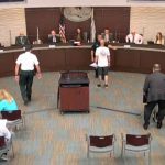 Mark Philips, in the white shirt to the right, is blocked by Sheriff's Cmdr. Phil Reynolds after Philips got in mayor Milissa Holland's face during this evening's council meeting. (© FlaglerLive via Palm Coast YouTube)