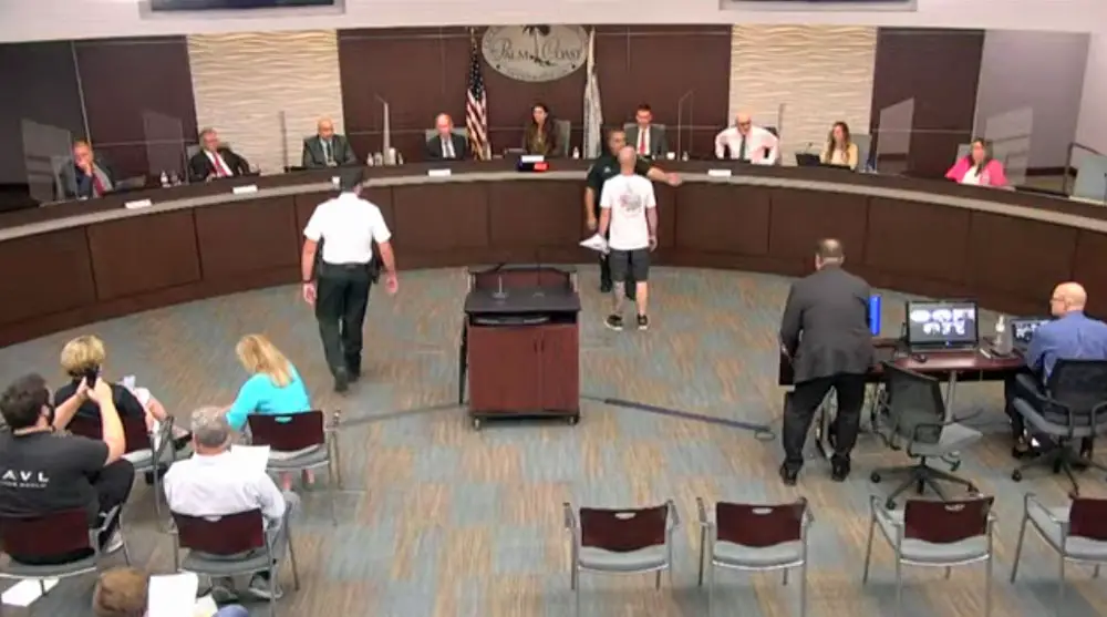 Mark Philips, in the white shirt to the right, is blocked by Sheriff's Cmdr. Phil Reynolds after Philips got in mayor Milissa Holland's face during this evening's council meeting. (© FlaglerLive via Palm Coast YouTube)