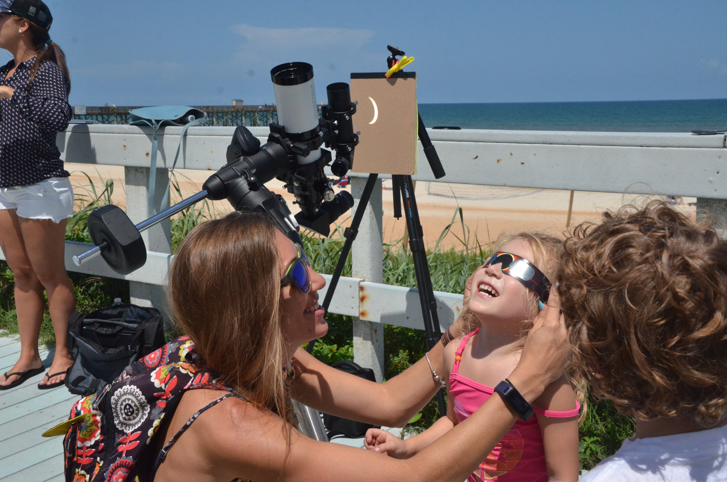 Kailani Raby, 3, of Flagler Beach, with her mother Tiffany Wiggen, catching the eclipse at its peak as it is also refracted by the telescope Scott Spradley had set up on the Flagler Beach Boardwalk this afternoon. Click on the image for larger view. (© FlaglerLive)