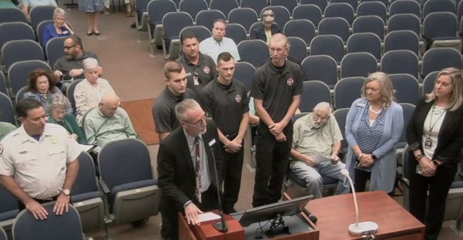 Superintendent Jim Tager, at the mic, and Fire Chief Don Petito, left, introduce Flagler County Fire Rescue's newest recruits this morning, and the first to be hired directly out of Flagler Palm Coast High School's three-year-old fire academy. The recruits are, from left, Noah Dunaway, Dylan Cronk and Beau Kruithoff, all 18. (© FlaglerLive via FCTV)