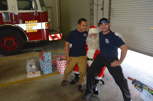 Firefighters need their Santa too: Lt. Armando Castaneda, left, and Fierfighter-Paramedic Matt Braasch sit on knees that in other times and places belong to Palm Coast firefighter Patrick Juliano. Click on the image for larger view. (© FlaglerLive)