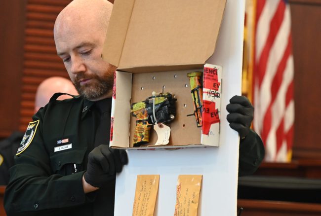 Flagler County Sheriff's deputy Joseph Oliva, one of the bailiffs in Circuit Judge Dawn Nichols's courtroom, shows the jury one of the three guns used in the fusillade that ended with Noah Smith's death in Bunnell in 2022. (© FlaglerLive)