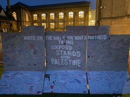 Visible in this image is an artist's recreation of the West Bank apartheid wall which guests of the encampment are invited to draw on. In the background are buildings belonging to Reuben College and the Bodleian Library's Radcliffe Science Library.
