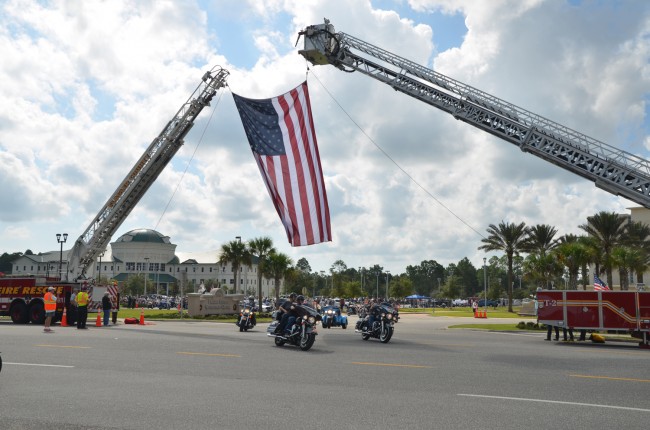 Riders streamed out of the Flagler County complex's parking lot under a giant flag draped by Flagler County Fuire Rescue and Palm Coast Fire Department personnel. Click on the image for larger view. (© FlaglerLive)