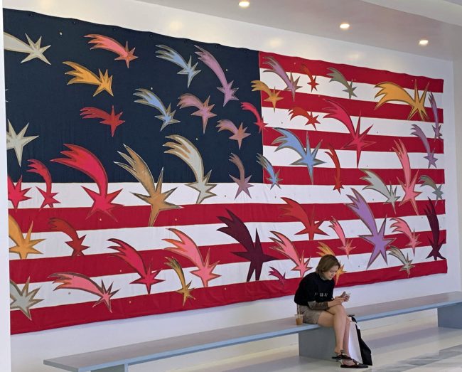 Friday is National Flag Day, which commemorates the adoption of the American flag on June 14, 1777. President Woodrow Wilson declared the first official Flag Day in 2016. Above, Frances Clemente's 'Wishing Stars,' a 2019 tapestry, hand-painted with natural dyes and stitched with embroidery and layered applique. The wooden pine bench on which the woman sits is part of the installation, currently at Hudson Yards, New York. The woman is not. Asked how he felt about sharing his work in that particular space, Clemente said: 'The audience of art is an audience of one. I hope he or she will pass by.' In that sense, the woman is very much a part of the installation. Click on the image for larger view. (© FlaglerLive)