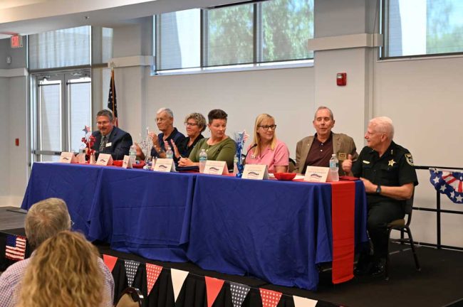 The panelists at today's Meet the Mayors event at FCAR. From left on the dais, County Commission Chair Andy Dance, Beverly Beach Mayor Steve Emmett, Flagler Beach Mayor Patti King, Marineland Mayor Angela TenBroeck, Bunnell Mayor Catherine Robinson, Palm Coast Mayor David Alfin, and Flagler County Sheriff's Chief Mark Strobridge. (© FlaglerLive)