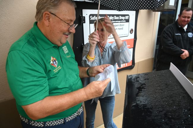 Bryant Perszyk, grand knight of the Knights of Columbus, trying to amke it through his speech before the rain ate it, with help from Council member Theresa Pontieri. Palm Coast Fire Chief Kyle Berryhill is to the right. (© FlaglerLive)