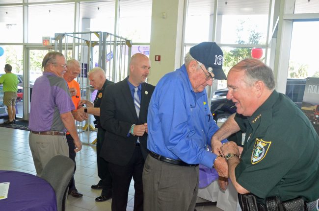 Sheriff Rick Staly takes pity on the men he's just arrested and starts undoing the handcuffs on County Commissioner Dave Sullivan, who'd be followed by Superintendent Jacob Oliva and County Commissioner Charlie Ericksen, who was being assisted by Chief Mark Strobridge. To the left is Steve Canfield, who's leading the Relay for Life fund-raising effort in the county this year, and who had dyed his mustache mauve for the occasion. (© FlaglerLive)