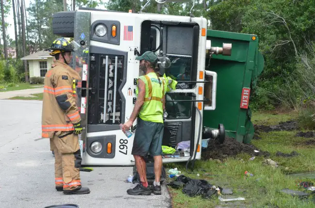A Palm Coast firefighters speaks with Bob Ackerman in front of the Waste Pro truck Ackerman, who was slightly injured, was driving when it overturned. Click on the image for larger view. (© FlaglerLive). 